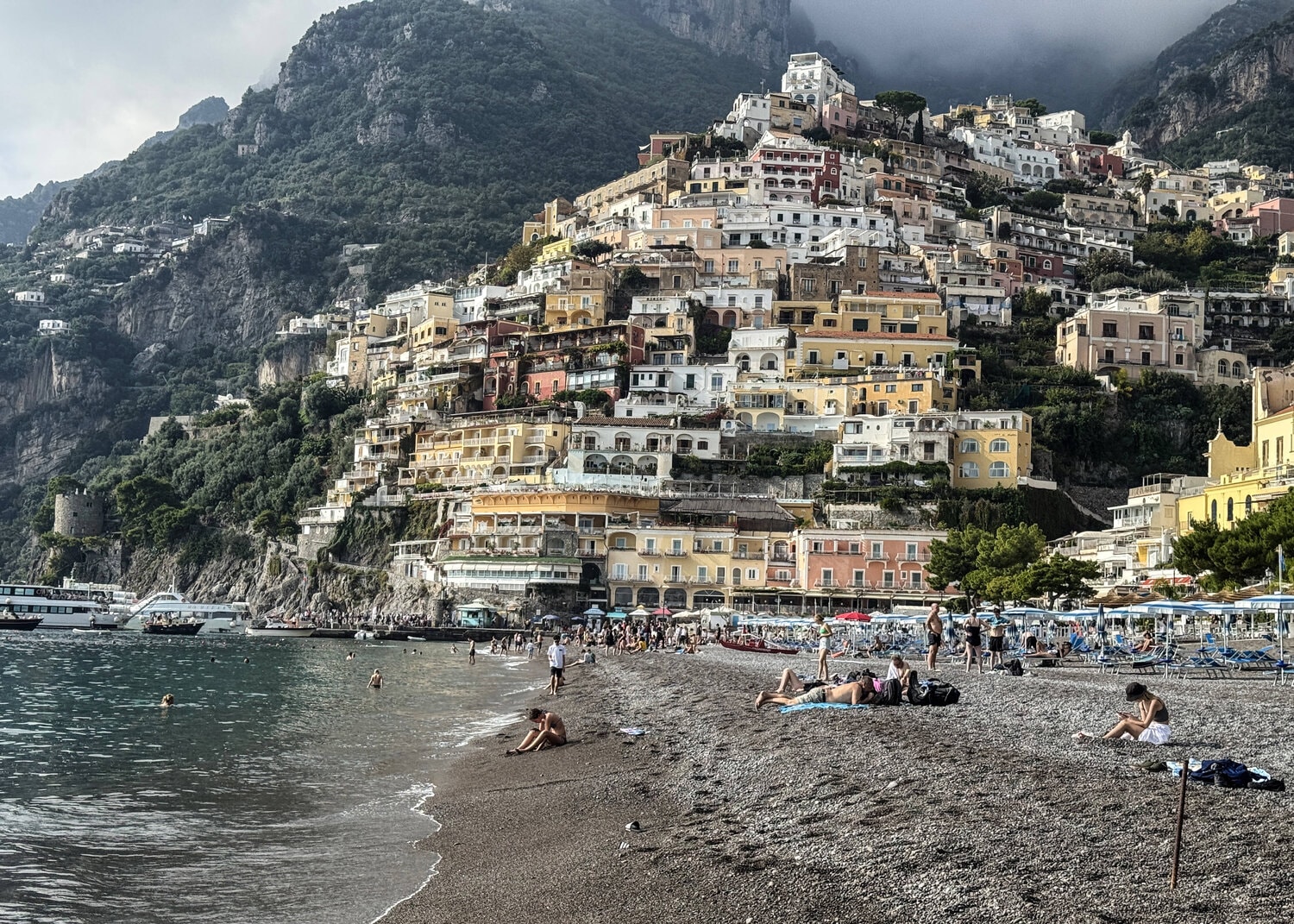 Blick auf den Strand von Positano mit zahlreichen Menschen, die auf dem Kiesstrand liegen, und den steil an den Berghang gebauten, pastellfarbenen Häusern.