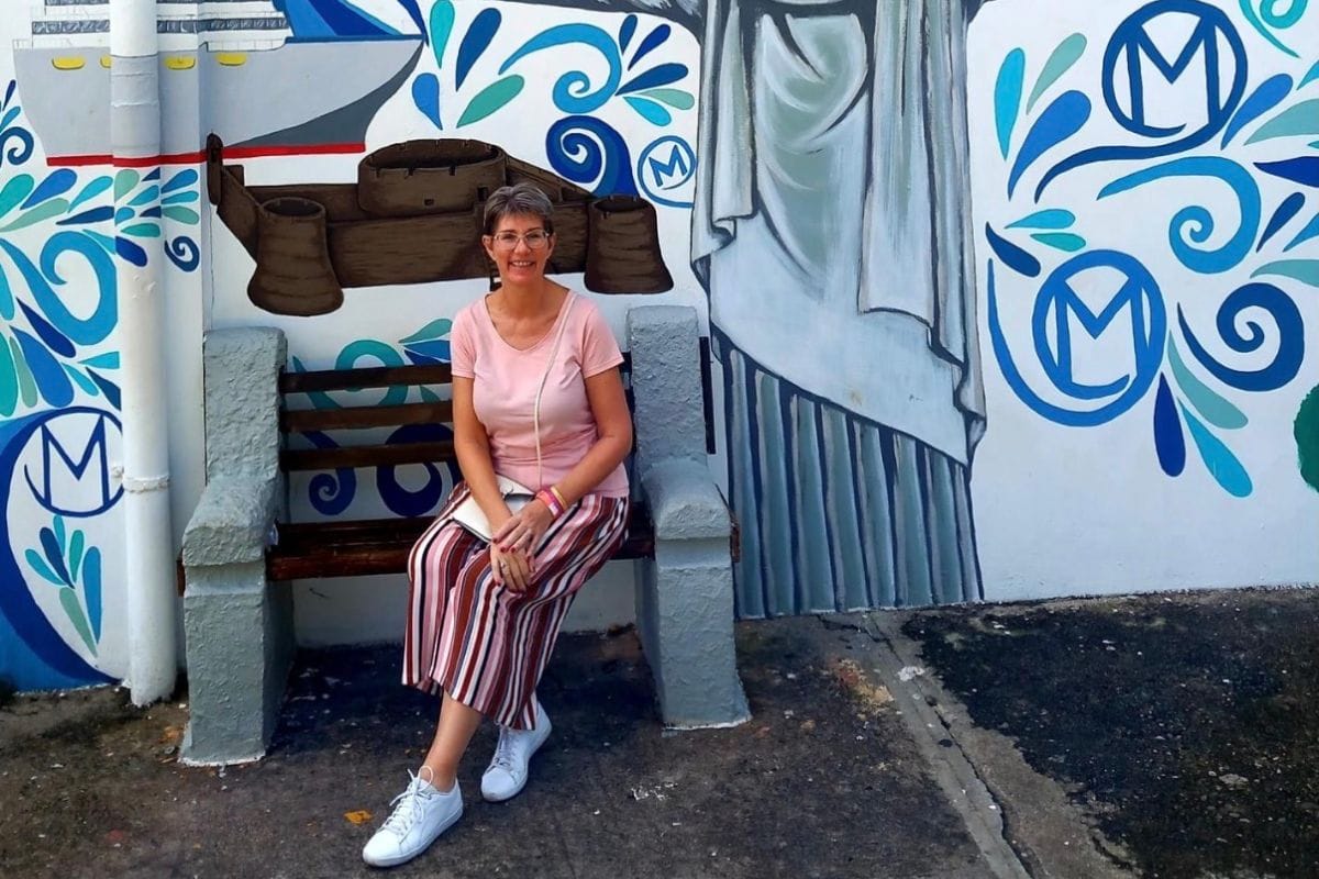 Eine Frau mit Brille sitzt auf einer Bank vor einer Wand mit einem großflächigen, blauen Wandgemälde, das eine Statue und maritime Symbole zeigt.