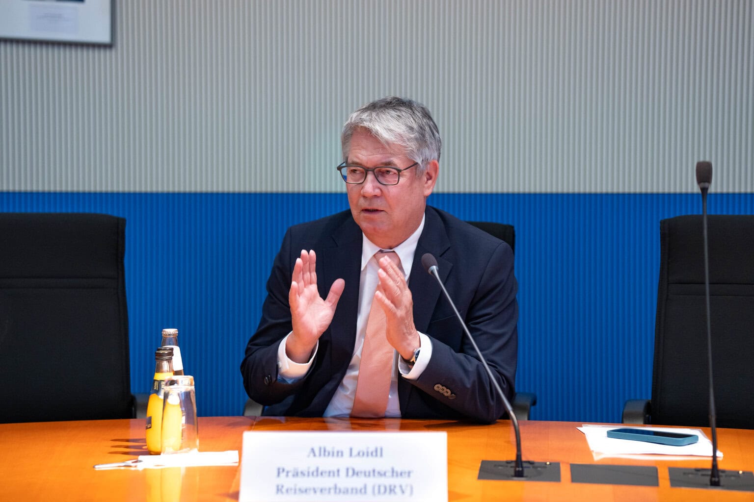 Ein Mann mit Brille und Anzug sitzt an einem Konferenztisch, gestikuliert mit den Händen und spricht in ein Mikrofon vor einem Namensschild mit DRV-Logo.