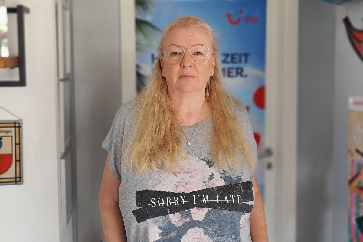 Eine blonde Frau mit Brille trägt ein graues T-Shirt mit Blumenmuster und dem Aufdruck Sorry I'm Late und steht vor einer Tür mit einem TUI-Werbeplakat.