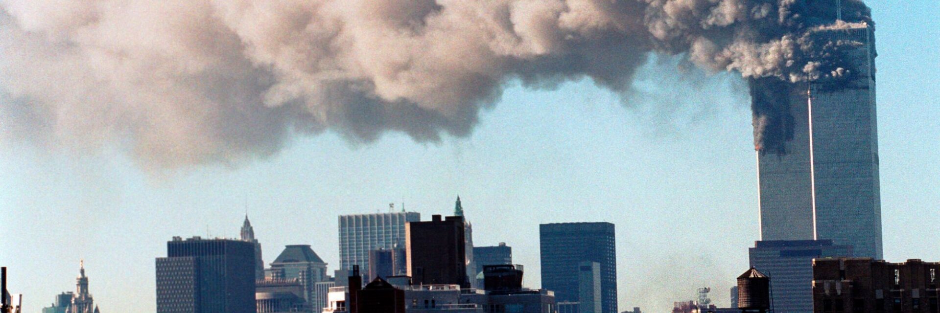 Blick auf die Skyline von New York City mit einem Wolkenkratzer, aus dessen oberem Bereich dichte, dunkle Rauchwolken in den hellen Himmel aufsteigen.