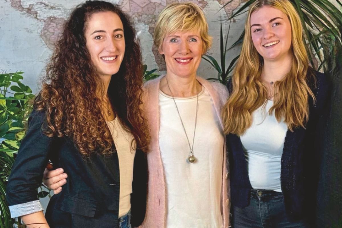 Drei lächelnde Frauen stehen nebeneinander vor einer Weltkarte an der Wand, flankiert von grünen Zimmerpflanzen in einem hellen Innenraum bei Tageslicht.