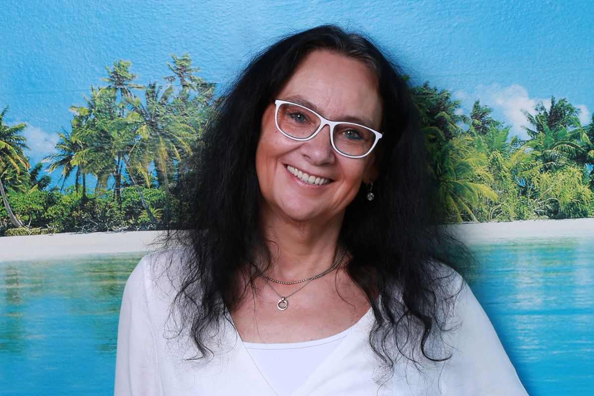 Eine Frau mit langem dunklen Haar und weißer Brille lächelt vor einem Hintergrund mit Palmen, Sandstrand und blauem Meer in die Kamera des Fotografen.