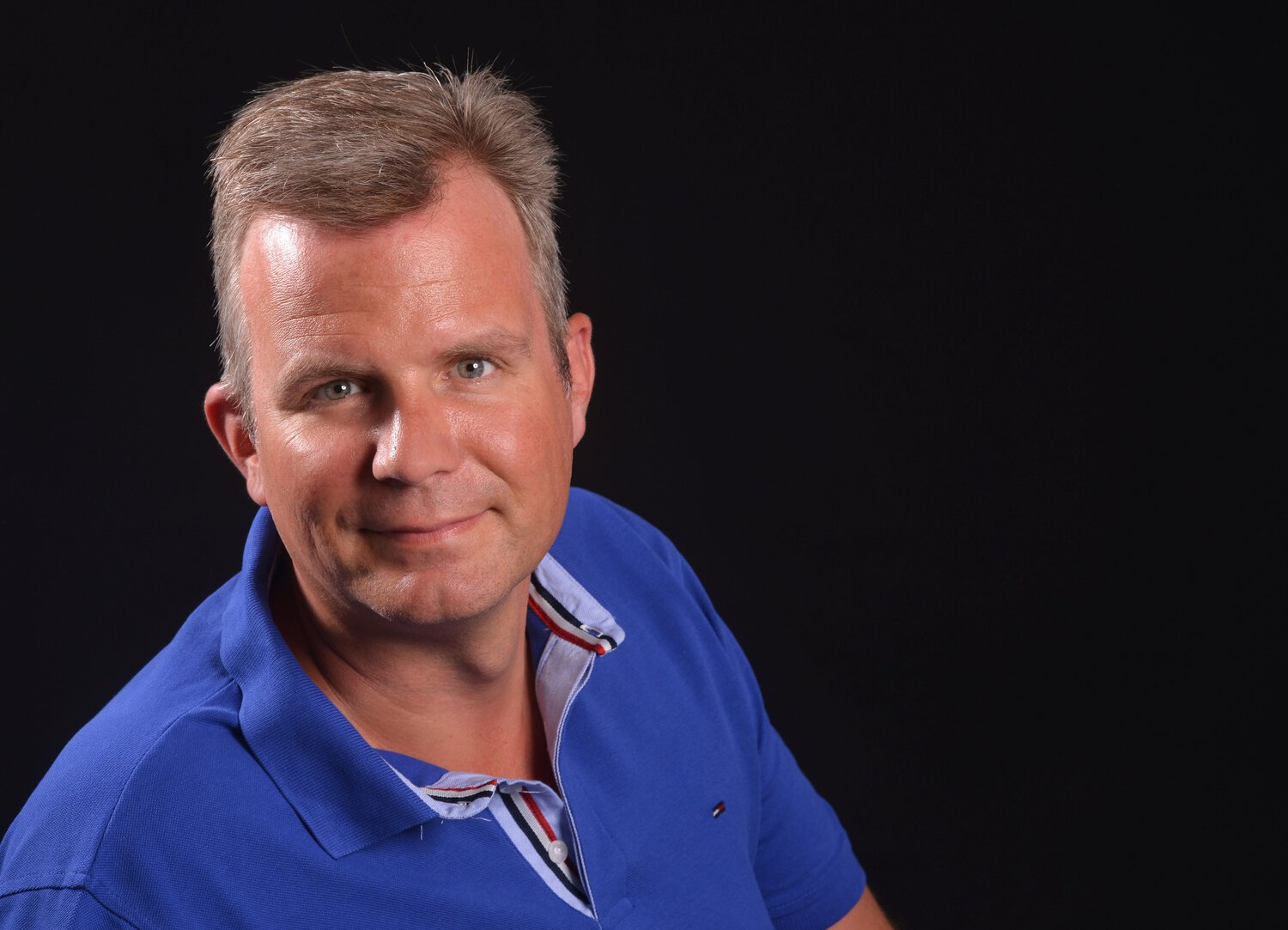 Ein Mann mit kurzem, hellem Haar und blauem Poloshirt blickt vor einem schwarzen Hintergrund direkt in die Kamera und lächelt leicht in die Linse hinein.
