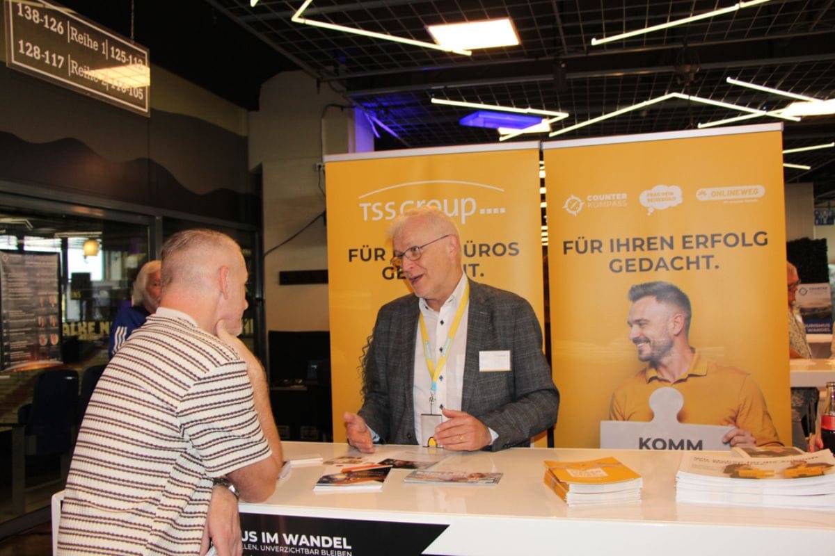 Ein Mann im Sakko spricht an einem Messestand mit einem anderen Mann, im Hintergrund stehen gelbe Roll-up-Banner mit dem Logo der tss group und Texten.