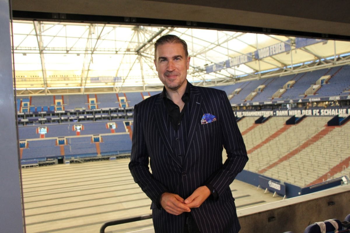 Ein Mann in einem dunkelblauen Nadelstreifenanzug steht in einer Loge mit Blick auf die leeren Tribünen eines großen Fußballstadions in Gelsenkirchen.