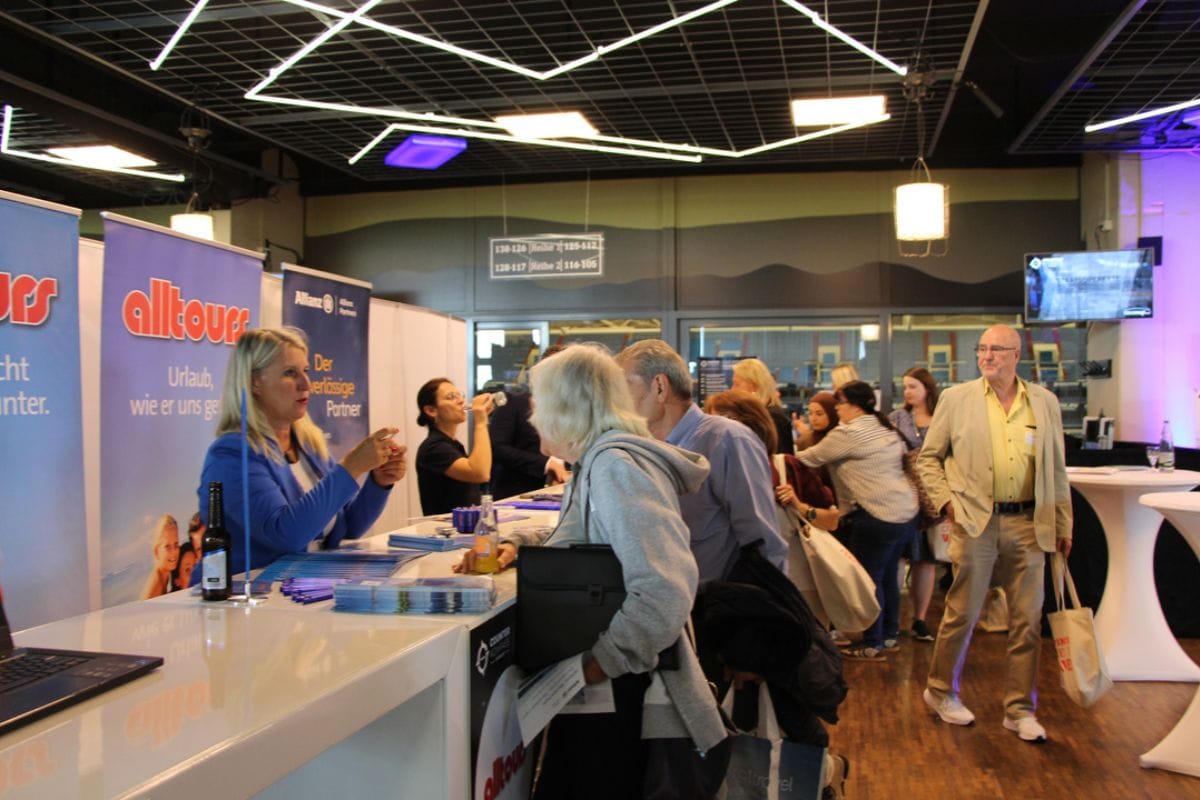 Eine Frau in blauem Blazer berät an einem Messestand von alltours Interessenten, während im Hintergrund weitere Personen in einem Konferenzraum stehen.