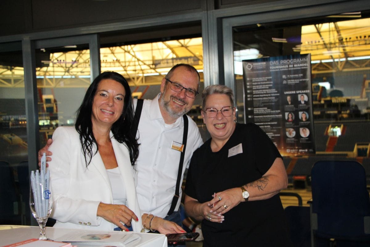 Drei Personen stehen nebeneinander vor einer Glasfront mit Blick in eine Arena; im Hintergrund ist ein Programm-Plakat mit der Überschrift „Das Programm“.