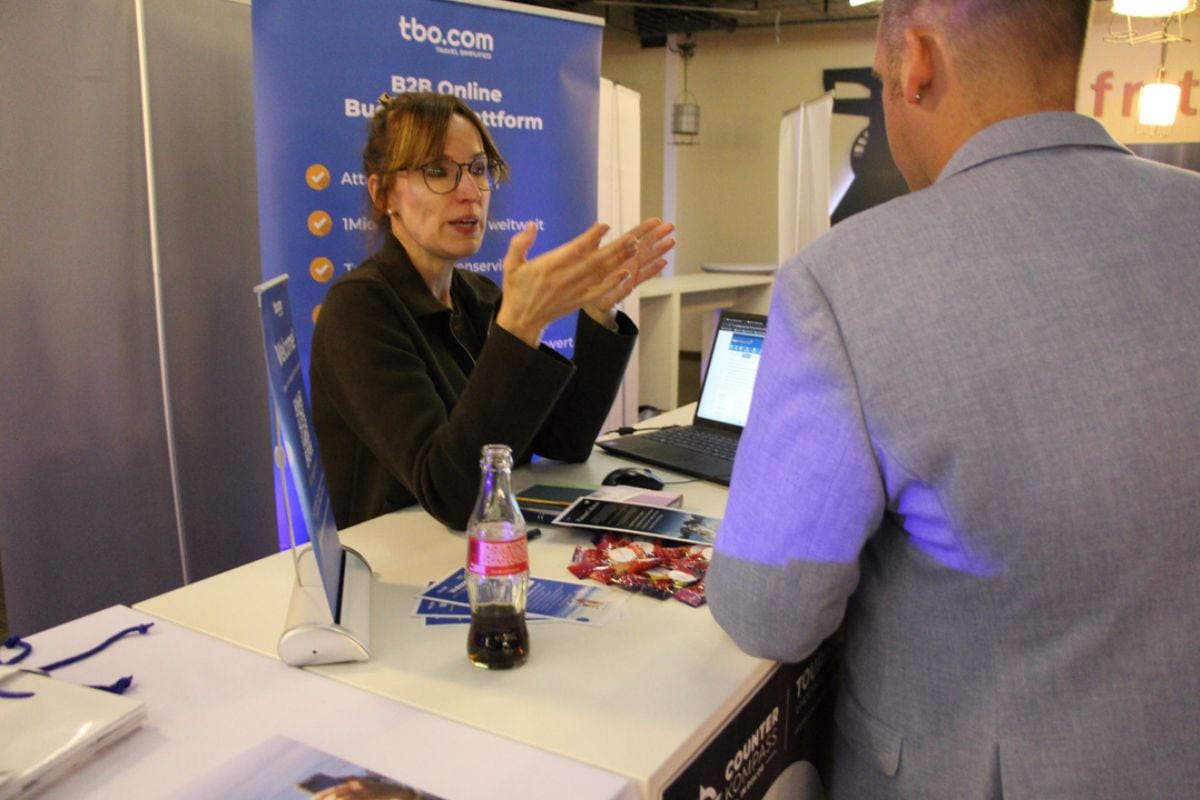 Eine Frau mit Brille spricht gestikulierend mit einem Mann an einem Messestand, auf dem ein Laptop, Werbematerialien und eine Glasflasche stehen.