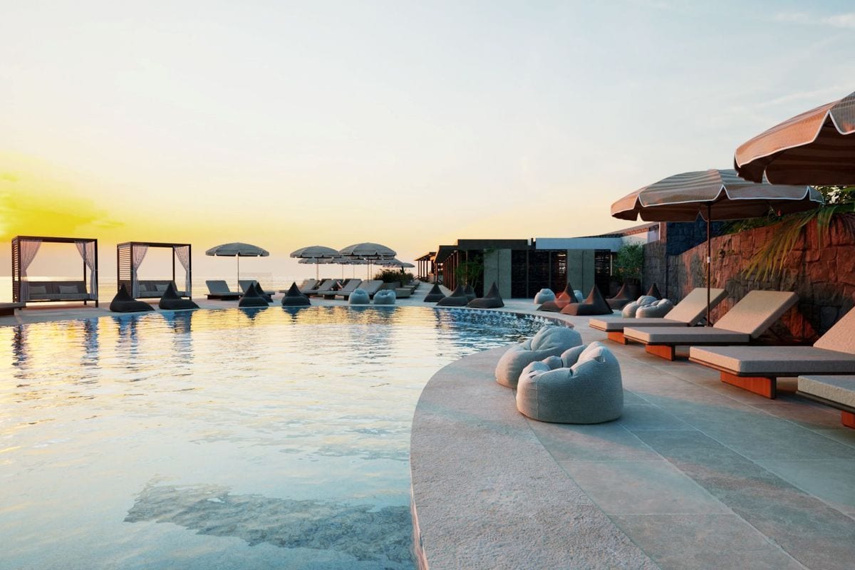 Ein Infinity-Pool bei Sonnenuntergang mit Liegen, Sonnenschirmen und Sitzsäcken auf einer weitläufigen Terrasse vor einer modernen Hotelanlage am Meer.