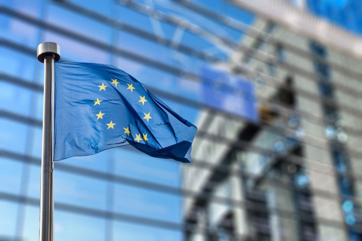 Die blaue Flagge der Europäischen Union mit ihren zwölf goldenen Sternen weht an einem Fahnenmast vor einer unscharfen Glasfassade eines modernen Gebäudes.