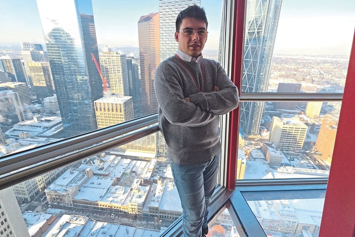 Ein junger Mann mit Brille und grauem Pullover steht vor einer hohen Fensterfront mit Blick auf eine verschneite Großstadt mit zahlreichen Hochhäusern.
