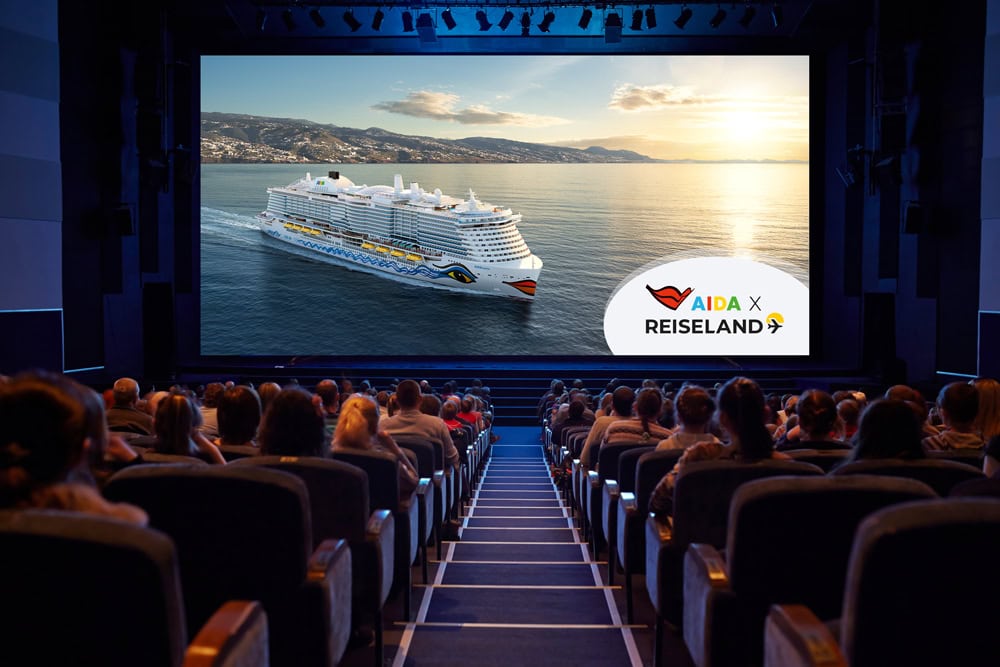 Blick in einen Kinosaal auf ein Publikum, das auf eine große Leinwand schaut, auf der ein AIDA-Kreuzfahrtschiff vor einer Küstenlandschaft zu sehen ist.