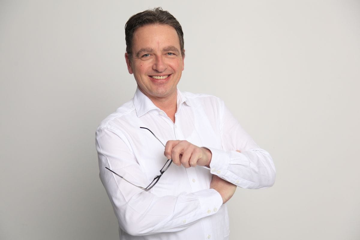 Ein freundlich lächelnder Mann mit kurzem braunem Haar trägt ein weißes Hemd und hält eine Brille in der Hand, vor einem neutralen, hellgrauen Hintergrund.