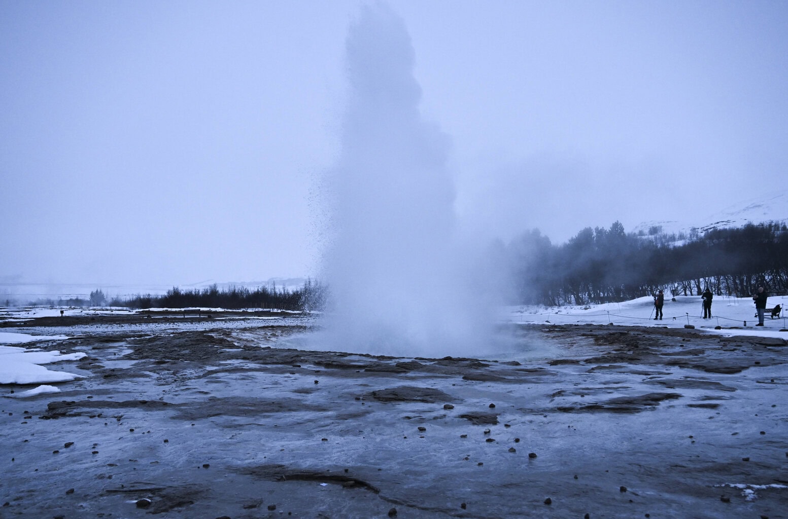 Eine hohe Fontäne aus heißem Wasser schießt aus einem Geysir in einer schneebedeckten Landschaft, während einige Personen das Naturschauspiel beobachten.