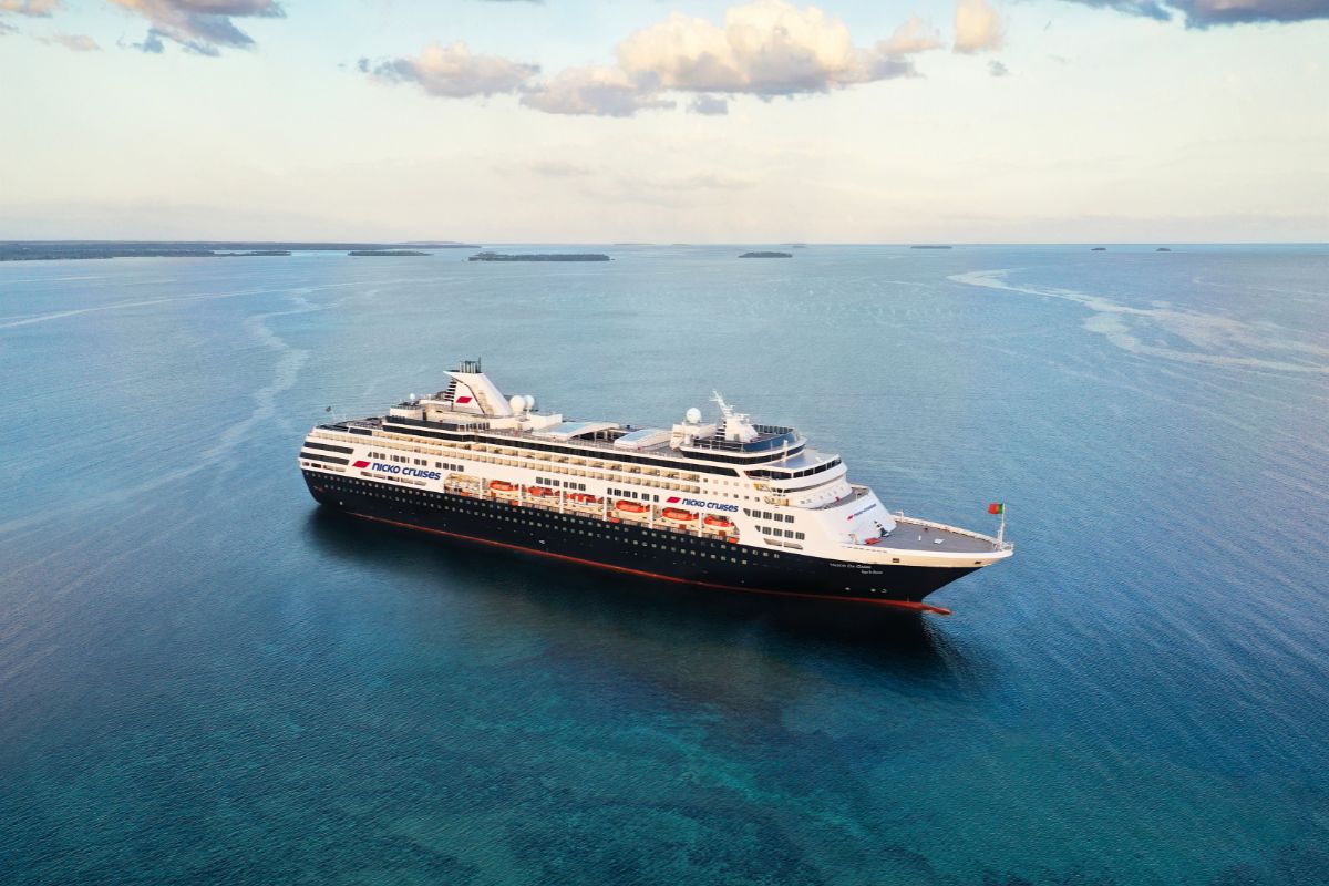 Ein Kreuzfahrtschiff der Marke nicko cruises fährt auf dem ruhigen, blauen Meer unter einem leicht bewölkten Himmel in einer weitläufigen Küstenlandschaft.