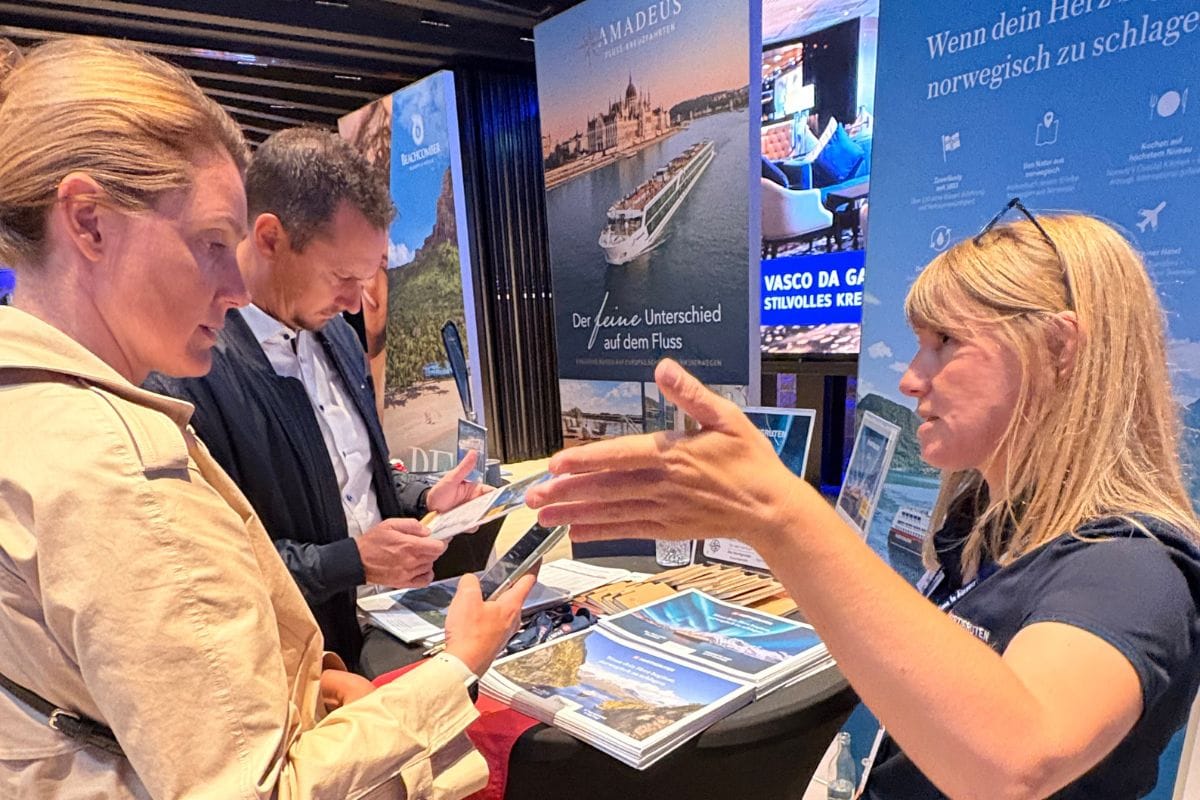 Drei Personen stehen an einem Messestand mit Infomaterial und Roll-up-Displays von Amadeus Flusskreuzfahrten und diskutieren miteinander im Innenraum.