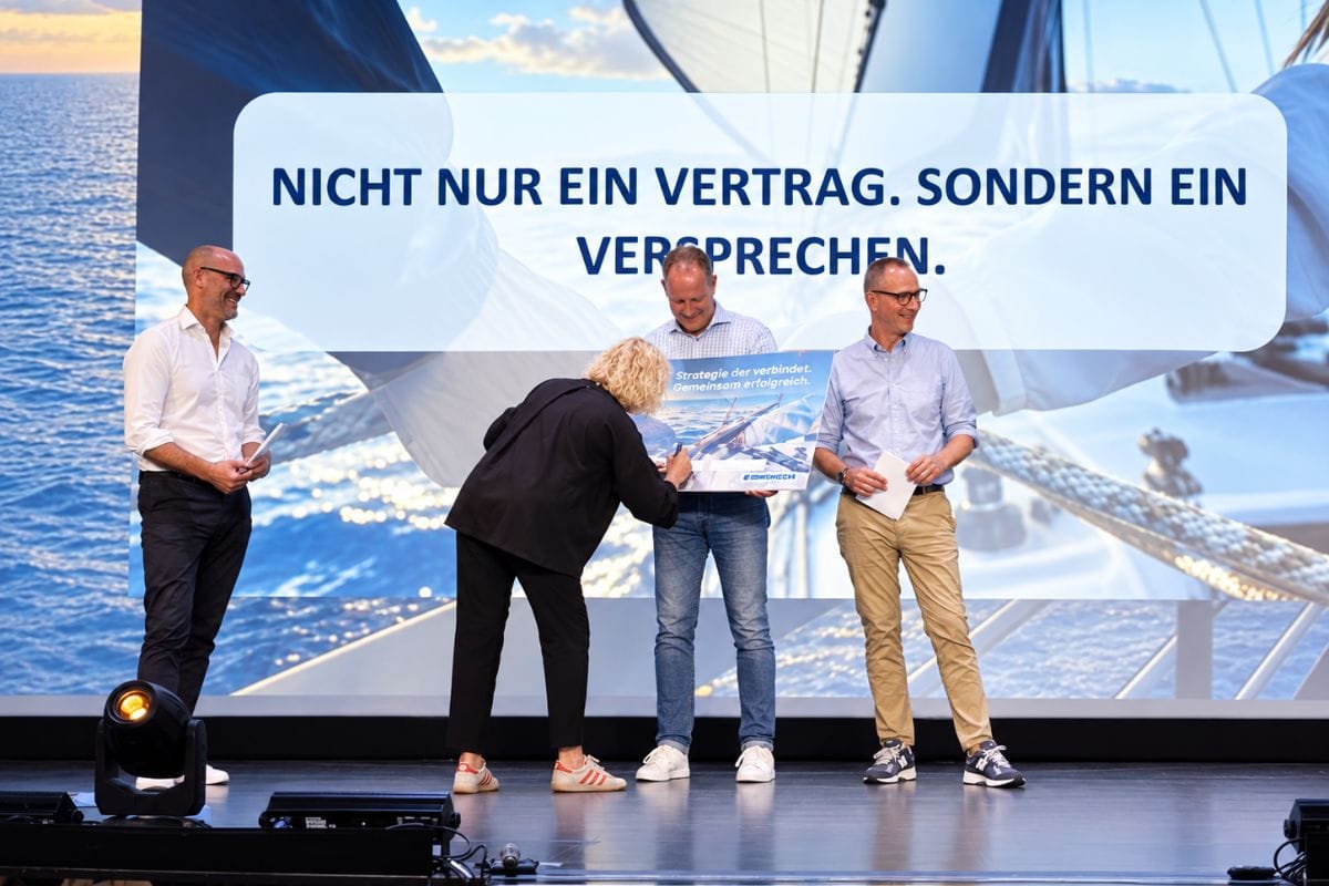 Drei Personen stehen auf einer Bühne vor einer Leinwand mit Segelboot-Motiv; eine Frau unterschreibt ein Schild, das von einem Mann gehalten wird.