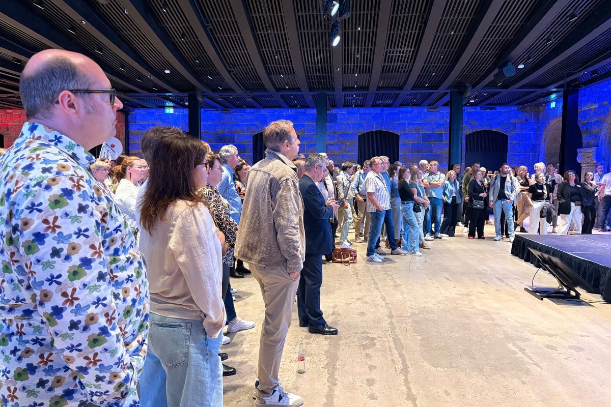 Eine Gruppe von Menschen steht in einem großen, modern beleuchteten Saal mit Steinwänden und blickt gemeinsam in Richtung einer Bühne im Hintergrund.