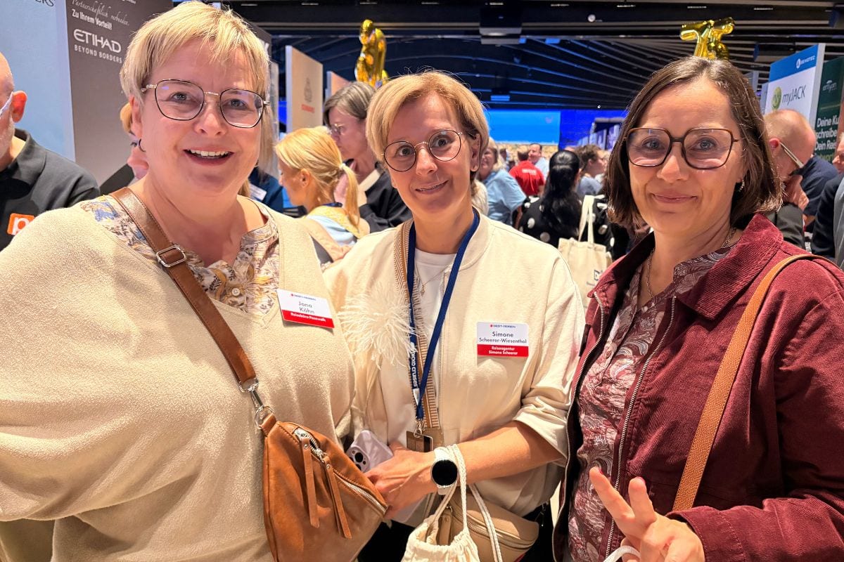 Drei Frauen mit Brillen stehen nebeneinander in einer Messehalle und lächeln in die Kamera, wobei zwei von ihnen Namensschilder an ihrer Kleidung tragen.