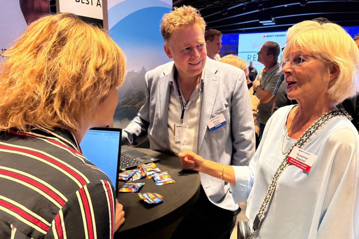 Drei Personen stehen an einem Stehtisch auf einer Messe. Eine Frau mit Brille und Namensschild spricht mit einem Mann im hellen Sakko und einer weiteren Frau.