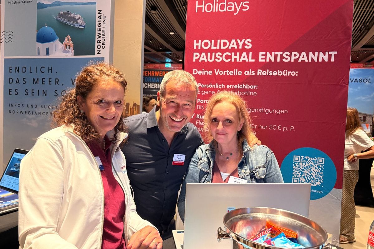 Drei Personen stehen lächelnd vor einem roten Werbebanner auf einer Messe, im Vordergrund steht ein Laptop und eine Schale mit Süßigkeiten auf dem Tisch.