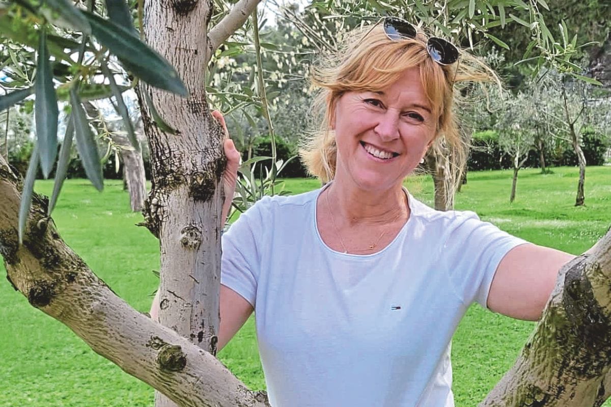 Eine lächelnde Frau mit blondem Haar und Sonnenbrille auf dem Kopf steht in einem Olivenhain zwischen den Ästen eines Baumes in einem weißen T-Shirt.