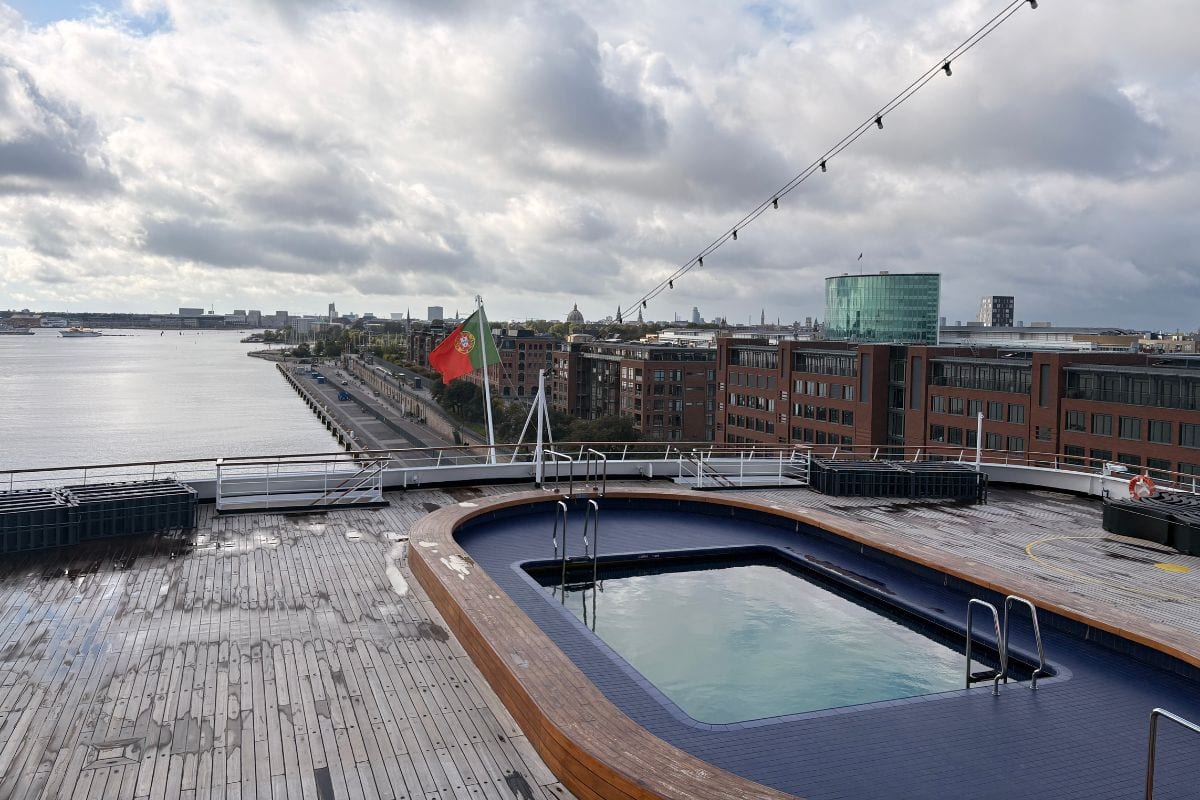 Blick von einem Kreuzfahrtschiffdeck auf einen Pool mit Holzumrandung, eine wehende portugiesische Flagge und die angrenzende Hafenstadt bei bewölktem Himmel.