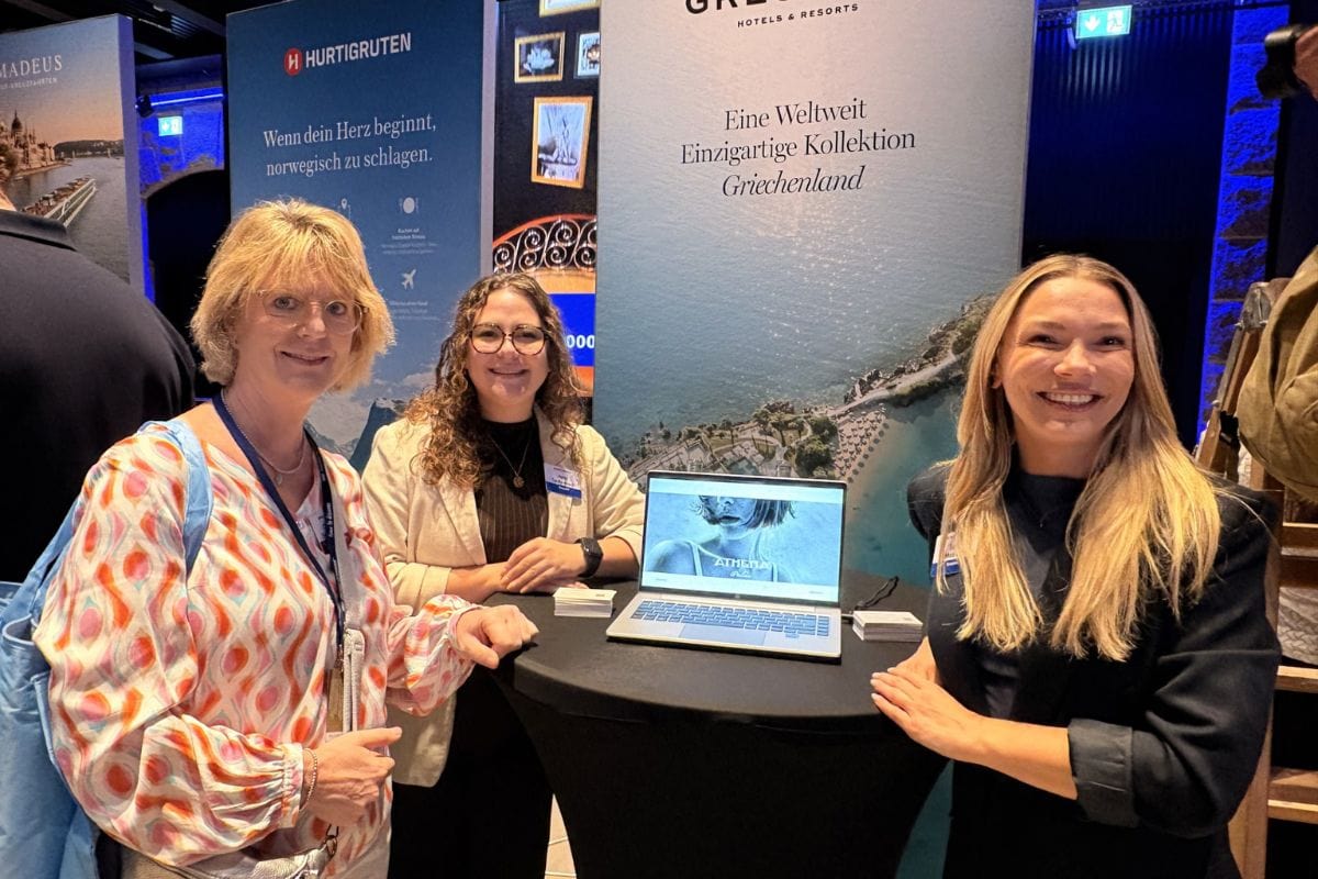 Drei lächelnde Frauen stehen an einem runden Stehtisch mit einem Laptop vor verschiedenen Werbeaufstellern von Hurtigruten und Grecotel Hotels & Resorts.