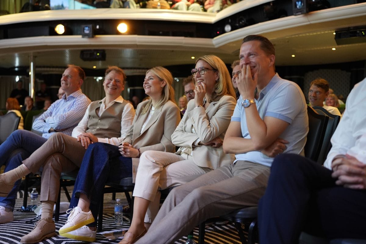 Eine Gruppe von Menschen sitzt in einem Theaterraum auf Stühlen und schaut in eine Richtung, während sie lächeln oder lachen. Sie tragen Business-Kleidung.