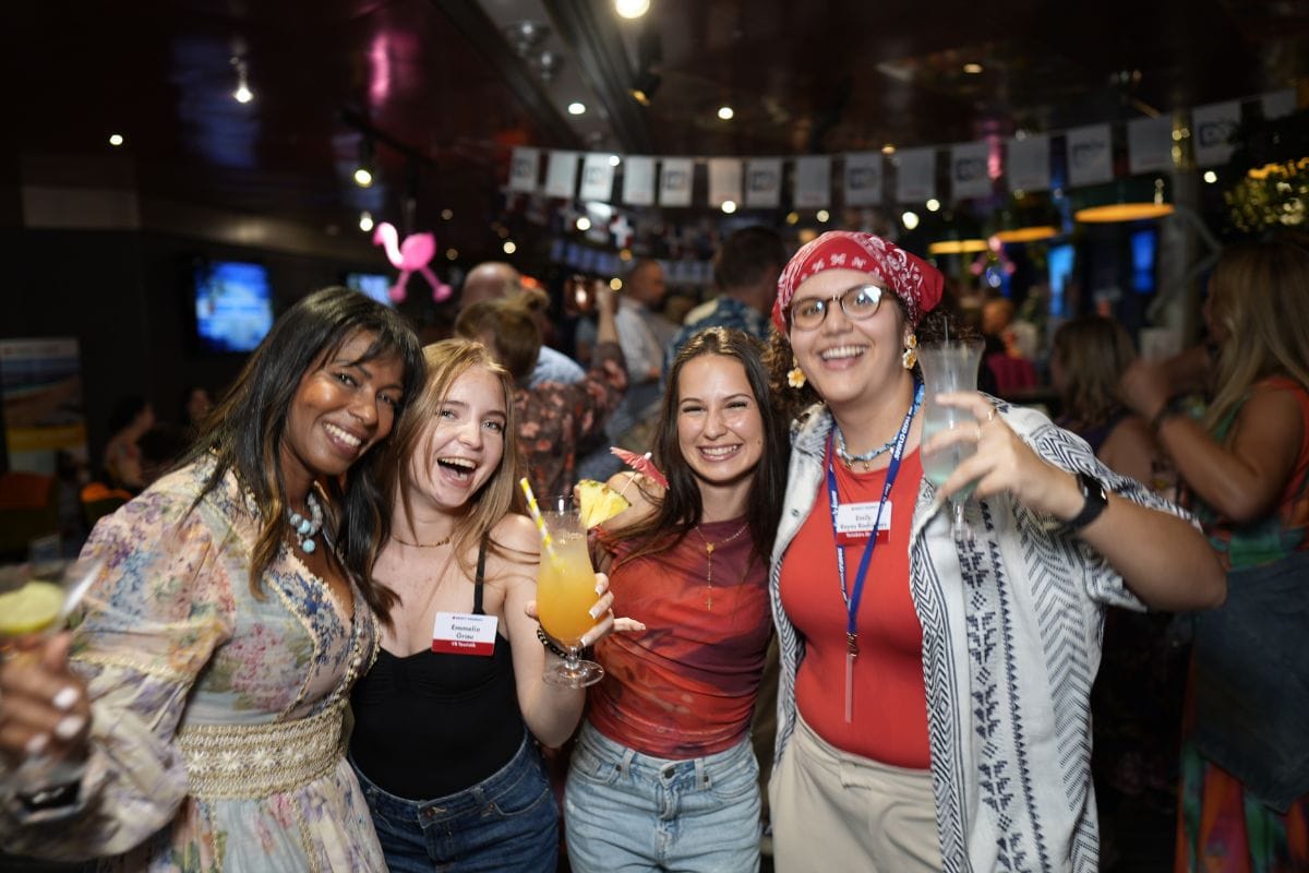 Vier junge Frauen stehen lächelnd in einer Bar zusammen, halten Cocktails in den Händen und tragen Namensschilder an ihrer Kleidung für eine Veranstaltung.
