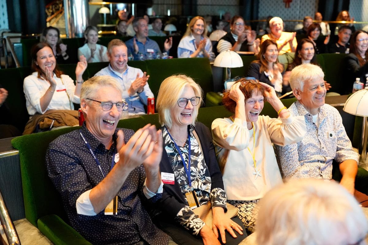Ein Publikum in einem Saal mit grünen Sesseln applaudiert und lacht herzlich, während einige Personen ihre Hände an den Kopf oder in die Luft halten.