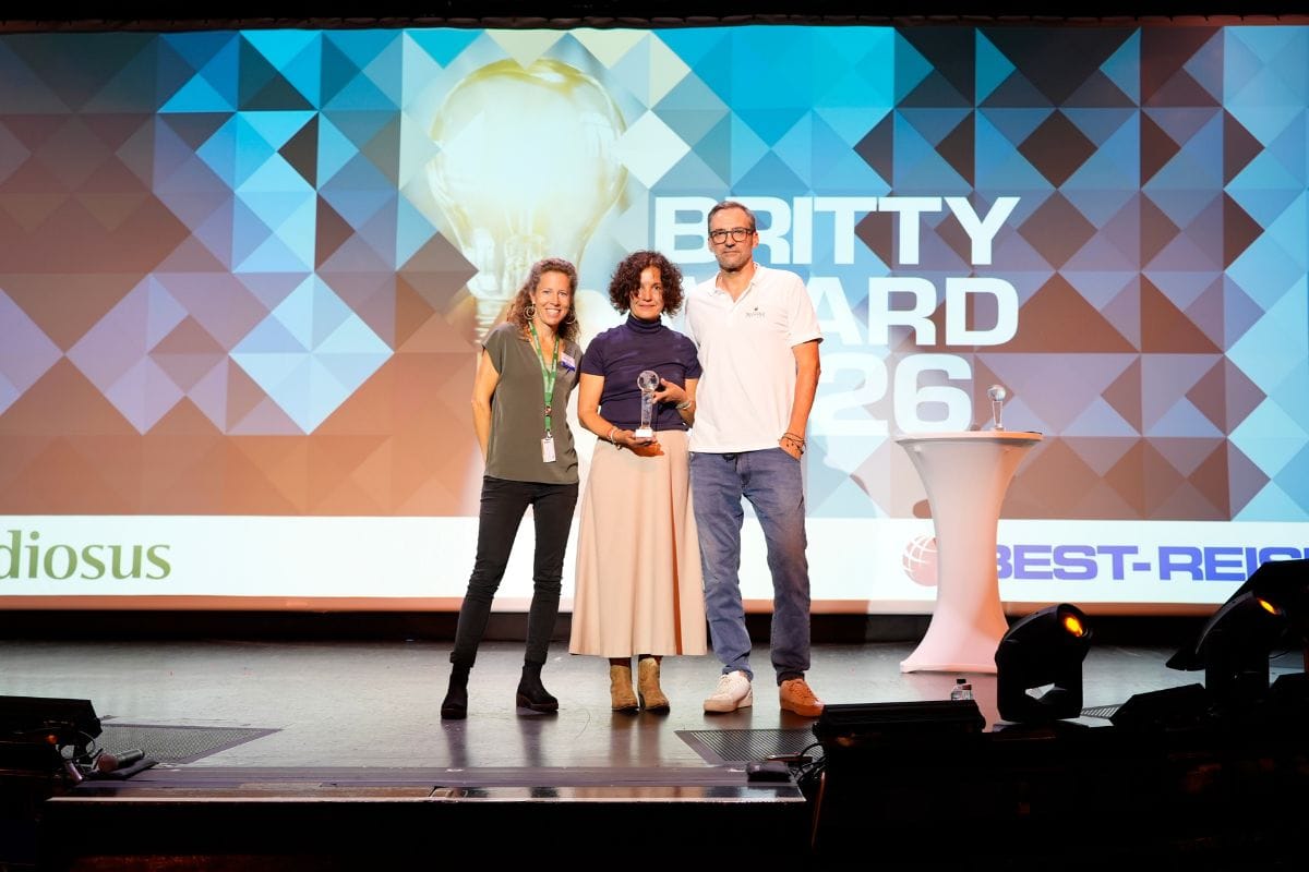 Zwei Frauen und ein Mann stehen auf einer Bühne vor einer Leinwand mit der Aufschrift BRITTY AWARD 26 und halten eine kleine gläserne Trophäe in Händen.