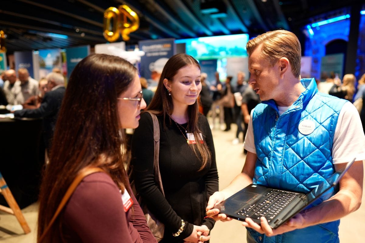 Ein Mann in einer blauen Weste hält einen Laptop und unterhält sich mit zwei jungen Frauen in einer belebten Veranstaltungshalle mit Messeständen im Hintergrund.