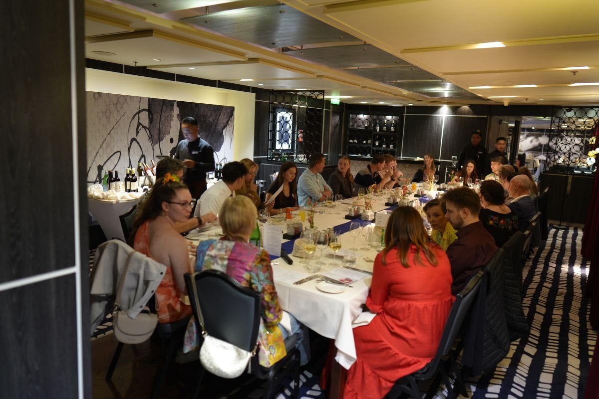 Eine Gruppe von Menschen sitzt an einer langen, weiß gedeckten Tafel in einem Restaurant mit dunkler Einrichtung und gestreiftem Teppichboden beim Essen.