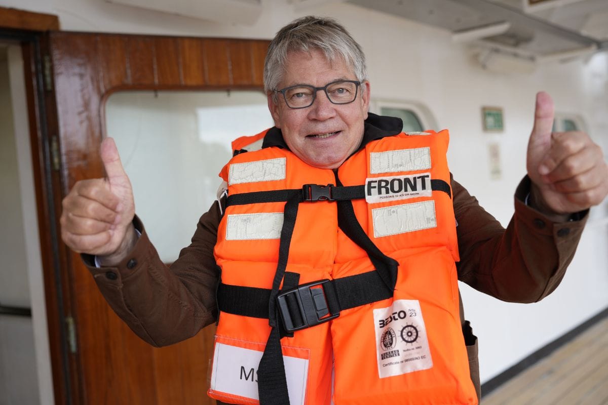 Ein älterer Mann mit Brille trägt eine leuchtend orangefarbene Rettungsweste über seiner braunen Jacke und zeigt mit beiden Händen den Daumen nach oben.