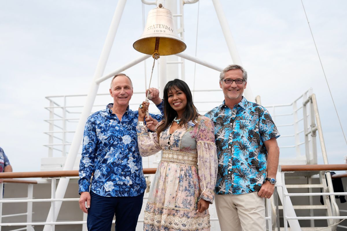 Zwei Männer in gemusterten Hemden und eine Frau in einem floralen Kleid stehen an Deck eines Schiffes unter einer Schiffsglocke mit der Aufschrift Statendam.