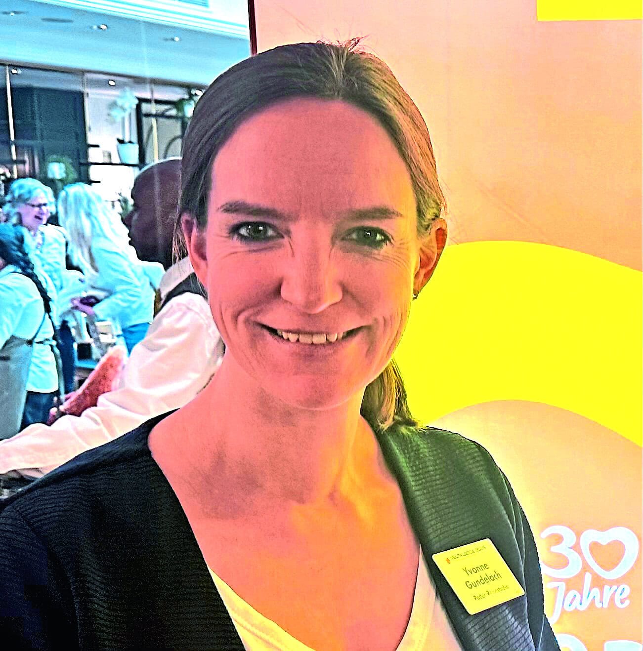 Eine Frau mit braunen Haaren und einem Namensschild am schwarzen Blazer lächelt in die Kamera, im Hintergrund sind verschwommene Personen und gelbe Flächen.