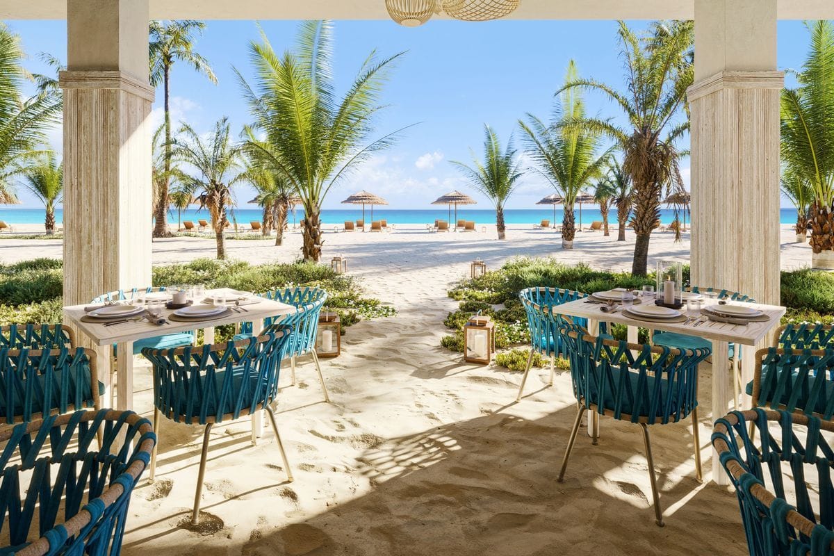 Blick von einer überdachten Terrasse mit gedeckten Tischen und blauen Stühlen auf einen weißen Sandstrand mit Palmen und das türkisfarbene Meer bei Tag.