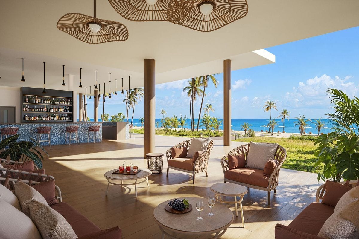 Eine offene Hotelbar mit Sitzgruppen auf einer Terrasse, die einen Blick auf den Strand, Palmen und das blaue Meer unter einem klaren Himmel bietet.