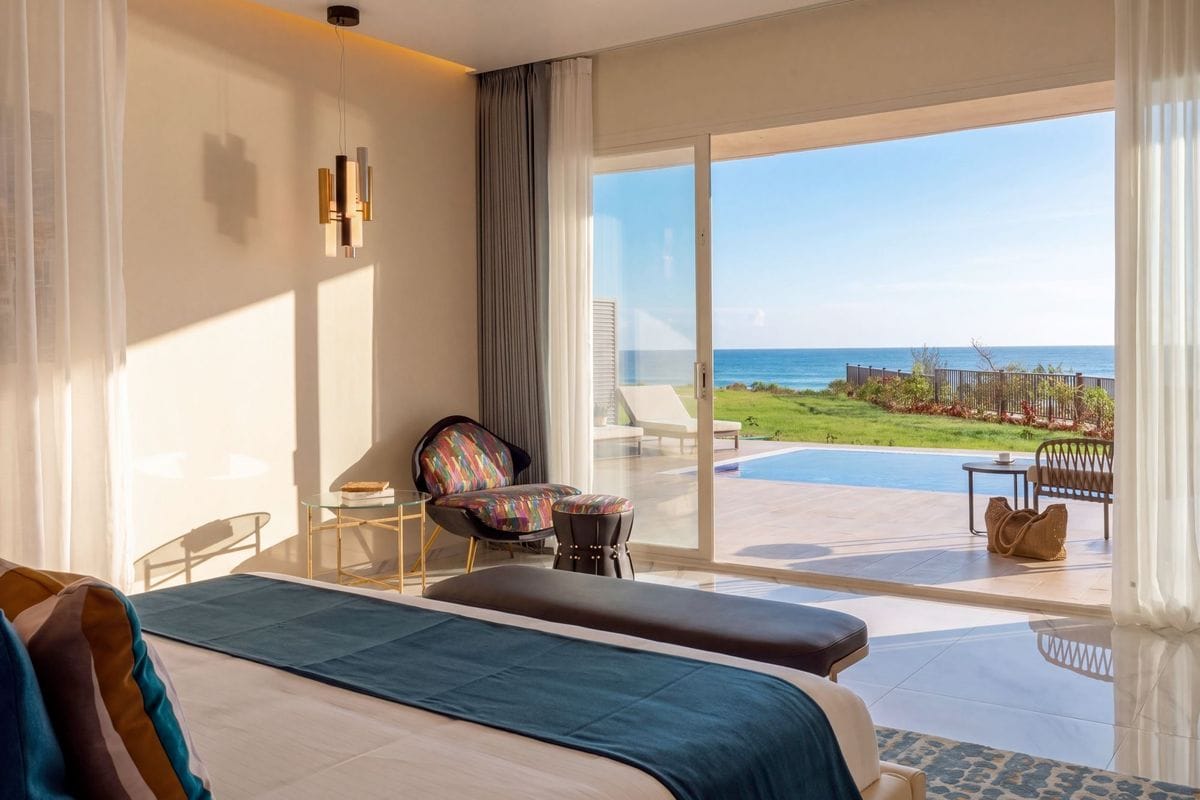 Ein helles Hotelzimmer mit einem Bett im Vordergrund und einer großen Schiebetür, die auf eine Terrasse mit privatem Pool und Meerblick hinausführt.
