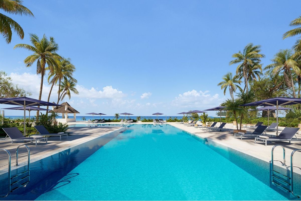 Ein langer, rechteckiger Swimmingpool mit türkisblauem Wasser, umgeben von Sonnenliegen, Sonnenschirmen und Palmen vor einem klaren, blauen Himmel am Meer.