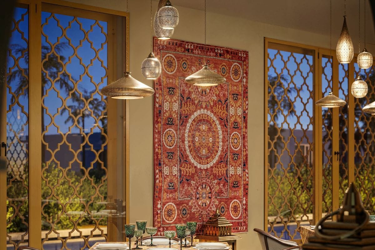 Ein Innenraum mit einem großen, gemusterten Wandteppich in Rot- und Goldtönen, beleuchtet durch mehrere dekorative Hängeleuchten vor hohen, verzierten Fenstern.