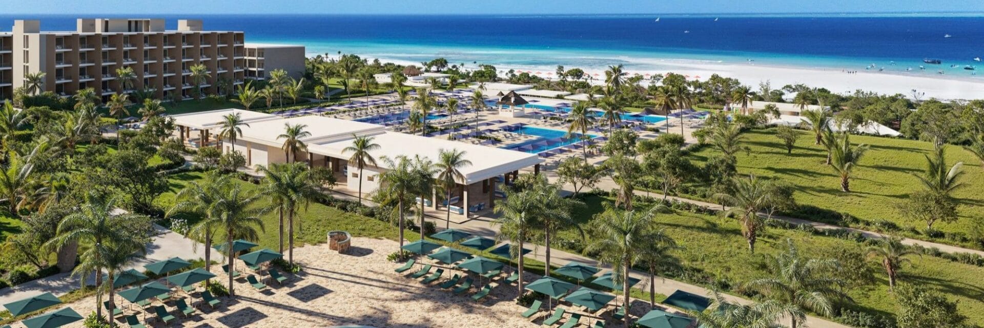 Iberostar_Selection_Zanzibar Luftaufnahme eines weitläufigen Hotelresorts mit mehreren Gebäuden, Poollandschaft, Palmen und Liegestühlen direkt an einem weißen Sandstrand am blauen Meer.