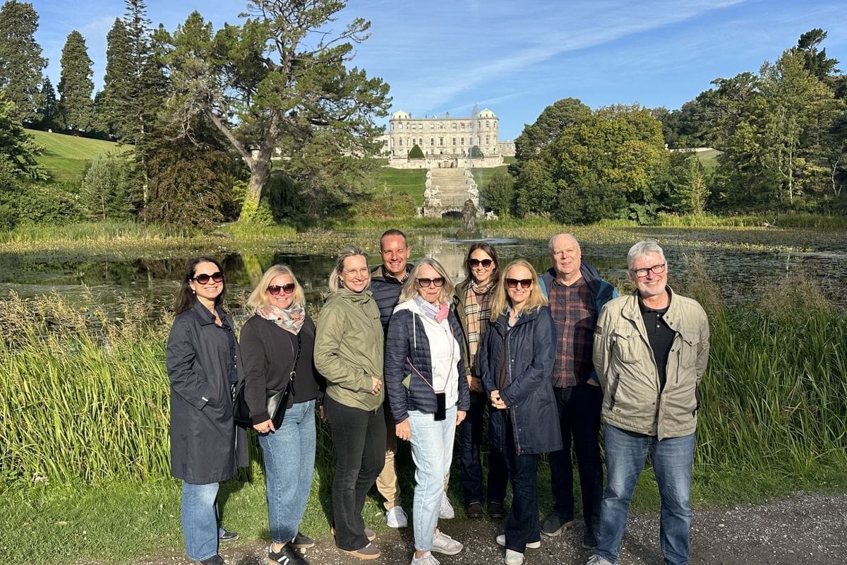 Eine Gruppe von neun Personen posiert lächelnd vor einem Teich und einer weitläufigen Parkanlage mit einem großen Herrenhaus im Hintergrund bei Sonnenschein.
