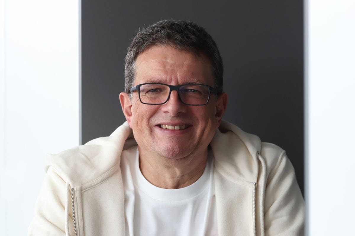 Ein Mann mit kurzem Haar und Brille lächelt freundlich in die Kamera. Er trägt ein weißes T-Shirt unter einer hellen Kapuzenjacke vor einem dunklen Hintergrund.