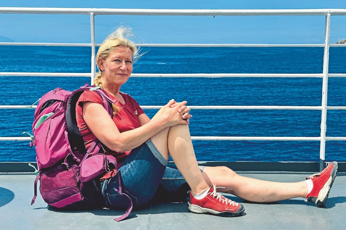 Eine blonde Frau mit einem pinken Rucksack sitzt auf dem Deck eines Schiffes vor einem weißen Geländer mit Blick auf das tiefblaue Meer bei Sonnenschein.