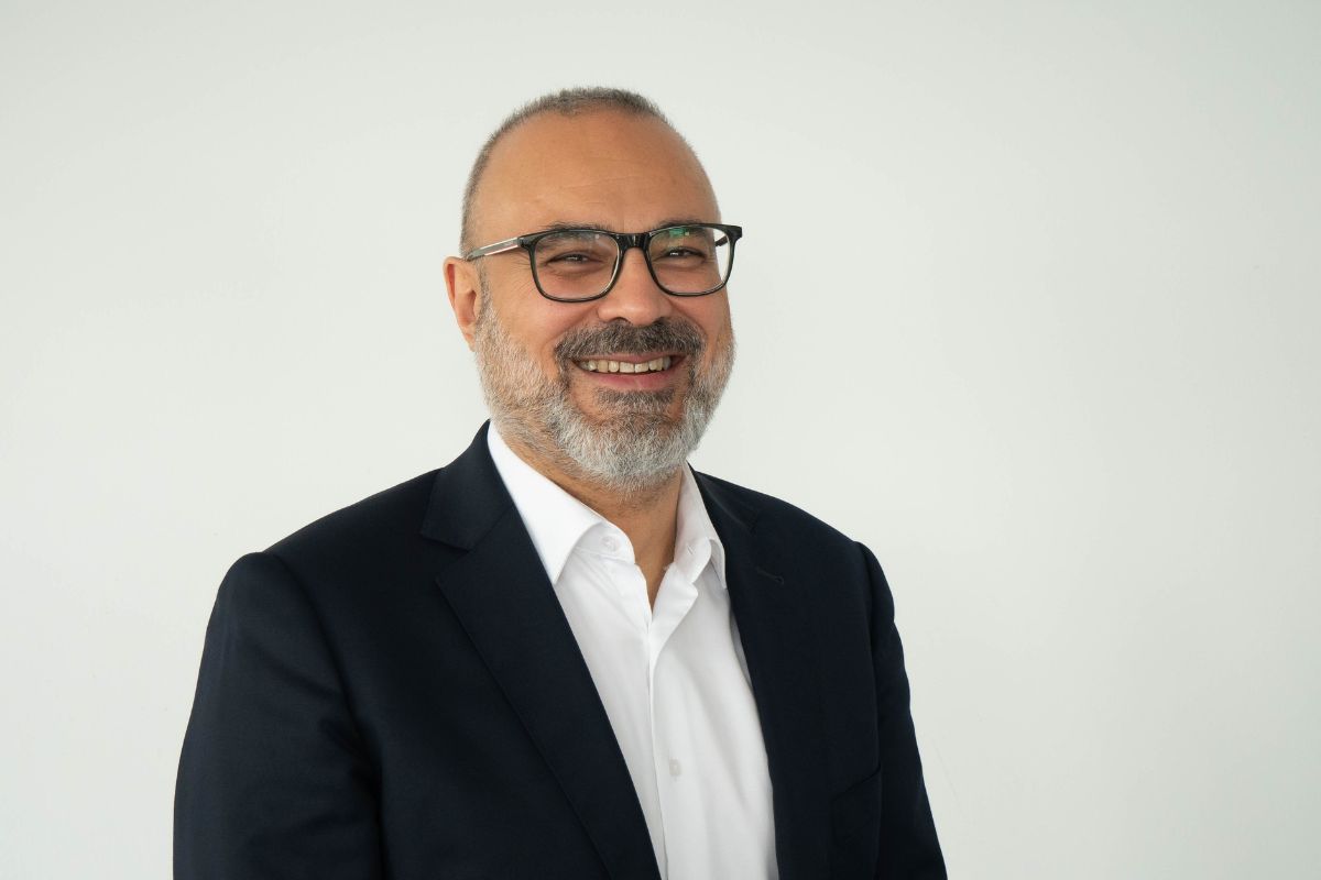 Ein lächelnder Mann mittleren Alters mit kurzem Haar, grau meliertem Bart und Brille trägt ein weißes Hemd unter einem dunkelblauen Sakko vor weißem Grund.