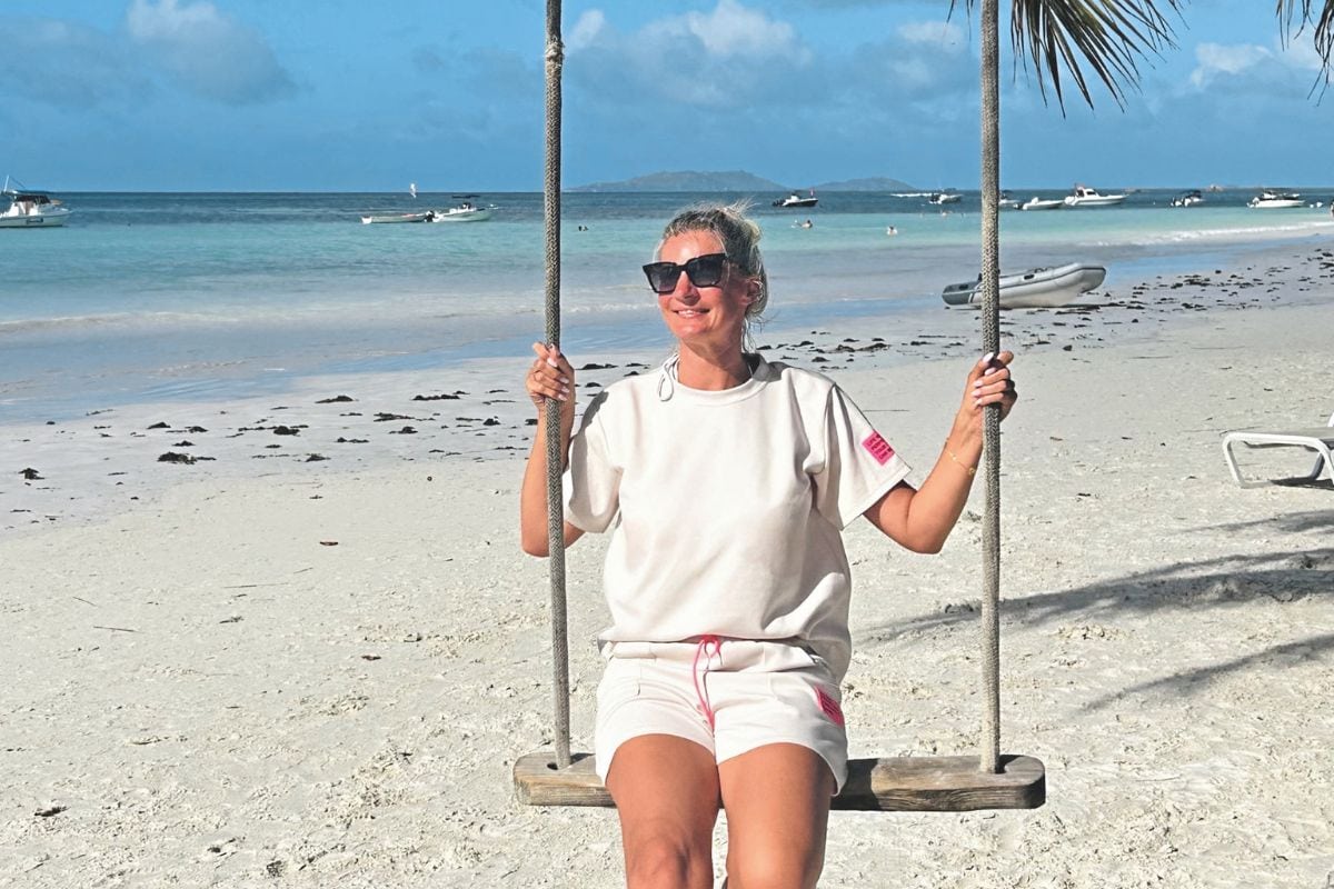 Eine Frau mit Sonnenbrille sitzt auf einer Holzschaukel an einem hellen Sandstrand, im Hintergrund ist das türkisfarbene Meer mit Booten zu sehen.