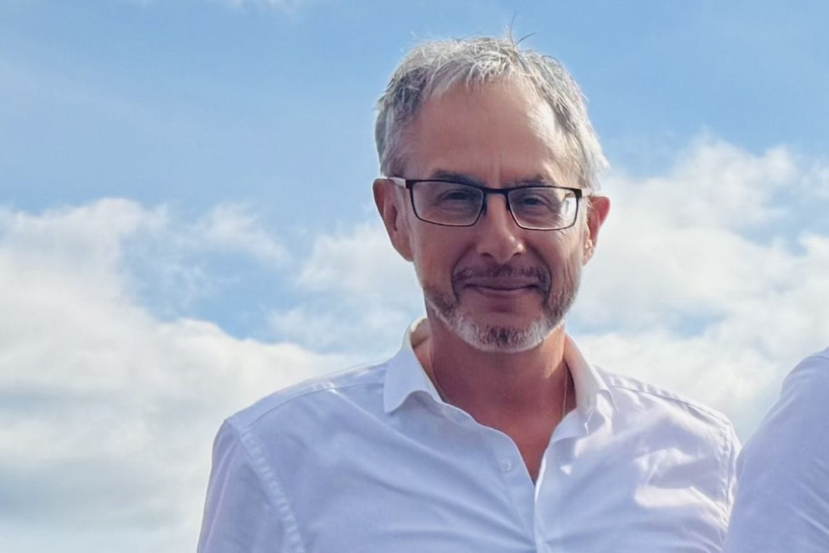 Ein Mann mittleren Alters mit graumeliertem Haar, Brille und einem weißen Hemd blickt vor einem hellblauen Himmel mit leichten Wolken in die Kamera.